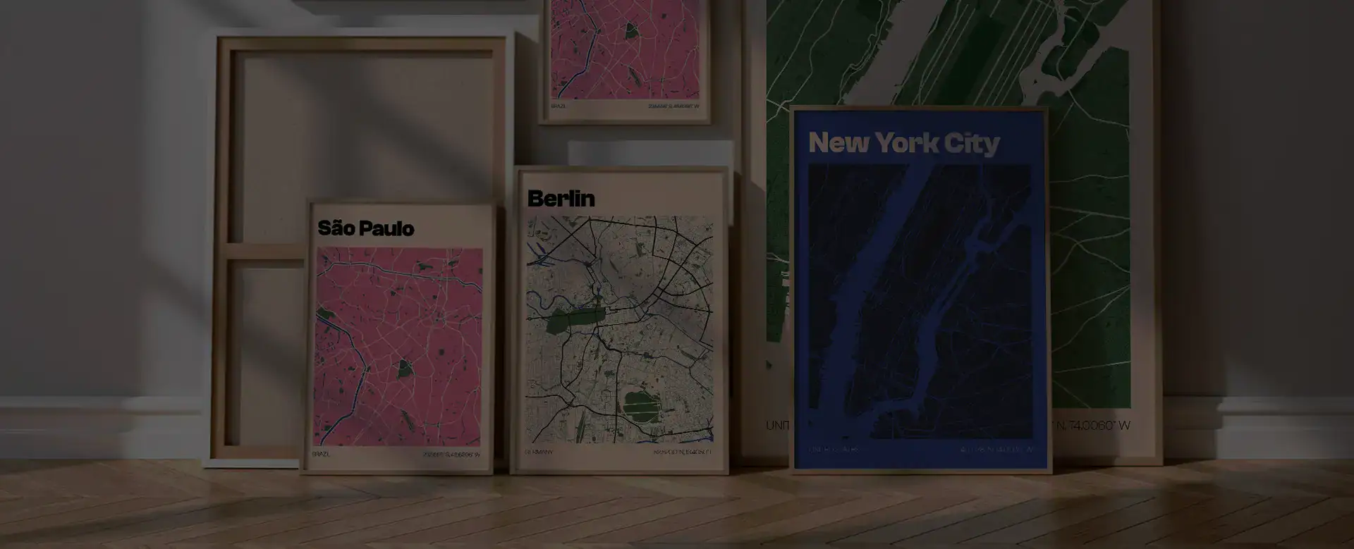 A collection of framed city map posters leaning against a wall, featuring maps of São Paulo, Berlin, and New York City with vibrant colors and modern designs, on a wooden floor.