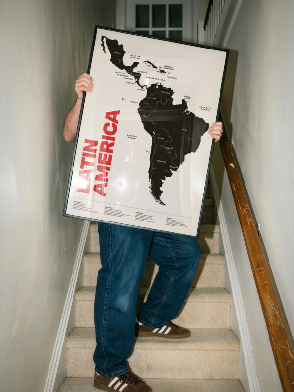 The Latin America Map Poster, a minimal geographic wall art print, features a detailed design of South and Central America in a black and white color scheme. It's displayed by a person on stairs, showcasing its modern aesthetic for home office decor.