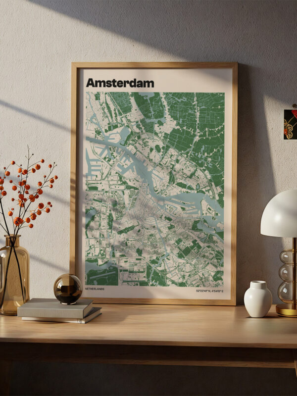 A framed map of Amsterdam displayed on a beige wall. The map features green areas representing the city's layout, with rivers and roads visible. A wooden table holds a decorative vase with orange berries, a lamp, and a small white object.