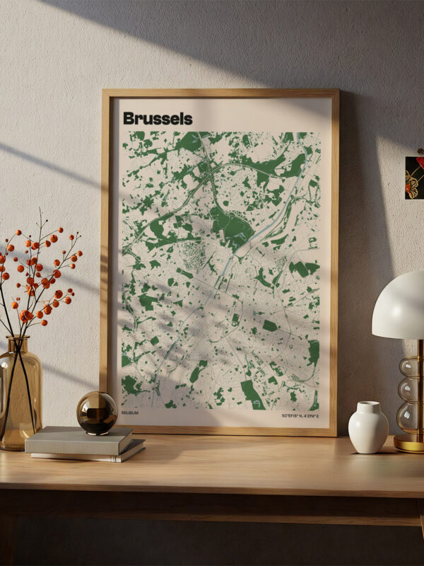A styled room featuring a wooden desk with a framed map of Brussels displayed prominently. The map, predominantly green on a light background, is complemented by decorative items like a lamp, a vase with branches, and a small bowl.