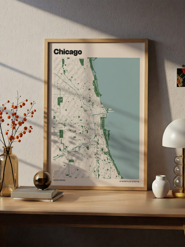 A framed map of Chicago is displayed on a wooden desk. The map is designed with green shades and outlines of streets. Nearby, there is a vase of orange berries, a black spherical object, and a lamp with a rounded shade, all casting shadows in natural light.