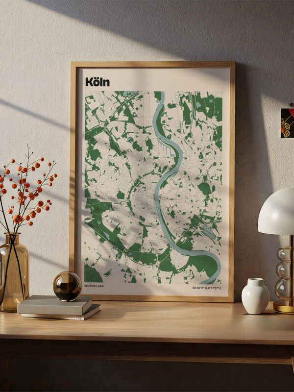A framed map of Köln displayed on a light wooden table. The map features a green and white color scheme with a river depicted. Nearby, there is a modern lamp, a small vase with orange berries, and a white decorative object.