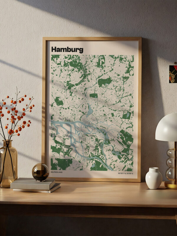 A framed map of Hamburg, displayed on a wall above a wooden desk. The map features green and white colors, with detailed outlines of streets and waterways. A vase with orange berries, a lamp, and a small decorative item are also on the desk.