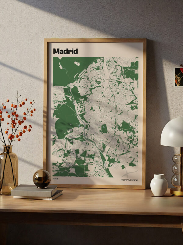 A framed map of Madrid displayed on a wooden desk near a decorative lamp and a vase with orange berries. The map features a contrasting green background with white outlines of the city streets.