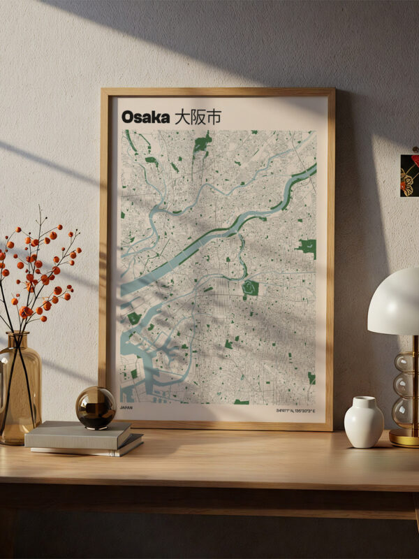 A framed map of Osaka displayed on a wooden desk. The map features a light-colored layout with streets and green spaces, labeled in English and Japanese. Nearby, a vase of orange berries, a decorative sphere, a lamp, and a small white vase are arranged on the desk.