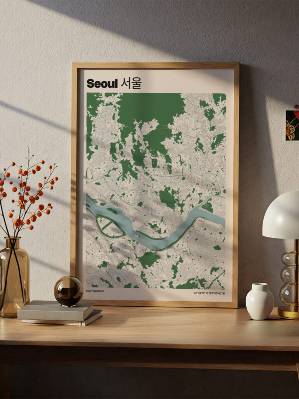 A framed map of Seoul displayed on a wooden table. The map features green areas and a river illustration. A vase with orange berries, a round black object, and a lamp are also on the table, with natural light casting shadows.
