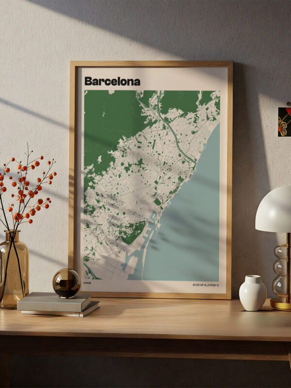 A framed map of Barcelona hangs on a wall, depicting the city in shades of green and gray. The map is displayed on a wooden table with a decorative vase of orange berries, a round black object, and a small lamp beside it.
